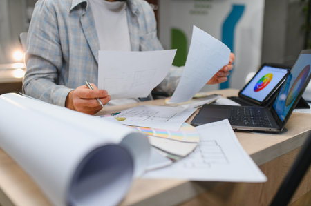 Interior designer working at desk, reviewing blueprints and plans. Laptop and tablet displaying...