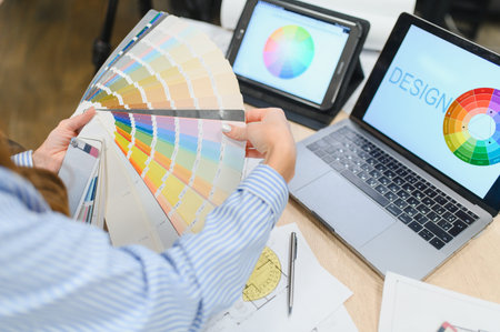 Designer selecting shades from a color palette fan, working on a laptop and tablet, creating visual...