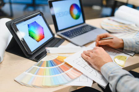 Architect or interior designer hands reviewing blueprints, using digital tablet with color palette...