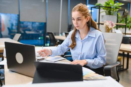 Young professional woman reviewing samples and taking notes while using a laptop in a contemporary...