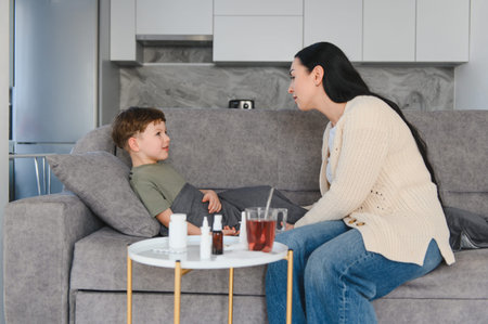 Mother sits beside sick young son on sofa, offering medicine and herbal tea while comforting him...