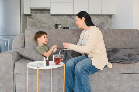 Loving mom giving medicine to her ill son resting on the sofa, providing care during his recovery at homeの写真素材
