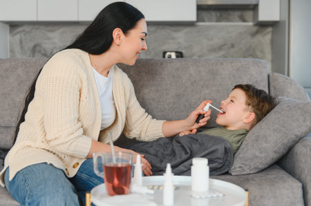 Mother applying throat spray to her sick boy relaxing on a sofa, providing home care for his sore...