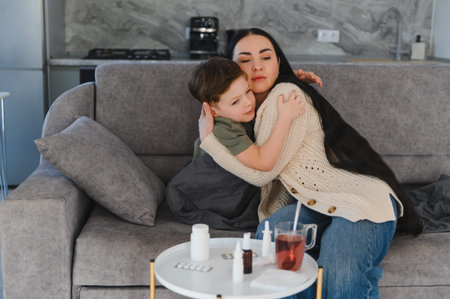 Mother embracing her unwell child on a couch at home, offering comfort while medicine and tea are on the tableの写真素材