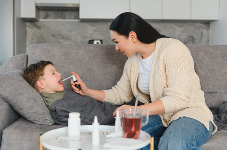 Mother treating her ill son with throat spray for a cough or flu symptoms while he rests on the sofa...