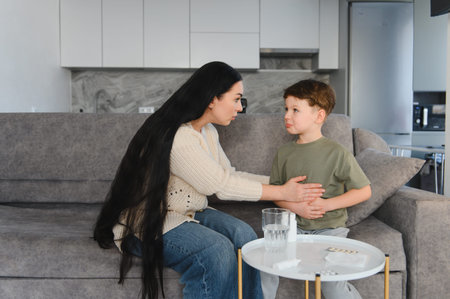 Mother consoling her little boy experiencing stomach pain while sitting on a sofa, with medicine on the tableの写真素材