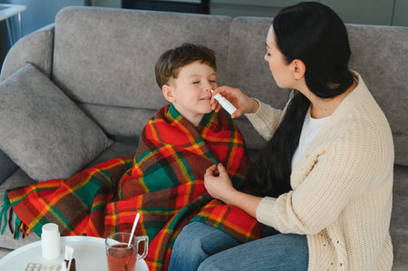 Mother applying nasal spray to her ill son wrapped in a blanket on the sofa, treating his cold or...