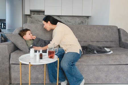 Mother comforting her ill child lying on a sofa at home. Table with medicine and a cup of tea nearby, highlighting health and careの写真素材