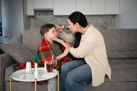 Mother checks her young sons temperature on the couch, offering blanket, medicine and comforting care during his illness at homeの写真素材
