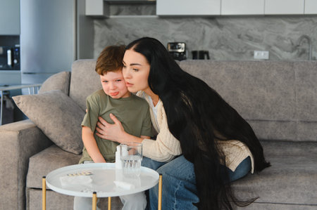 Mother consoling her crying child feeling unwell, sitting on a sofa with medicine and a glass of water nearbyの写真素材