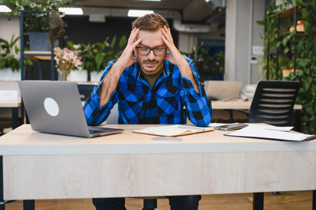 Man feeling stressed and exhausted from work, holding his head while sitting at a desk with a laptop...