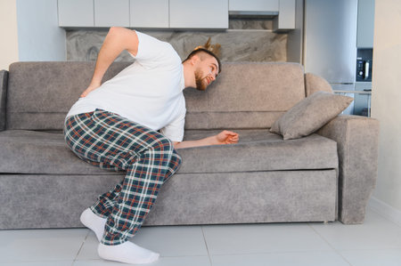 Young man experiencing severe low back pain at home, sitting on sofa and holding his painful lower...