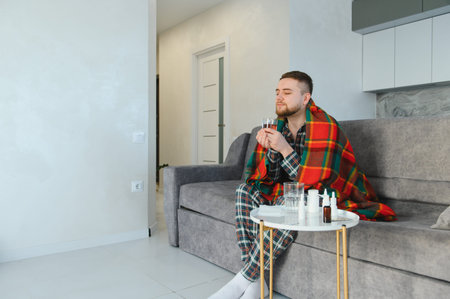 Man feeling unwell and sick with a seasonal illness, sitting on a sofa wrapped in a blanket and drinking warm teaの写真素材