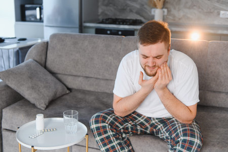 Man feeling dental pain, holding his cheek, needing medication on the sofa at home for oral health discomfortの写真素材