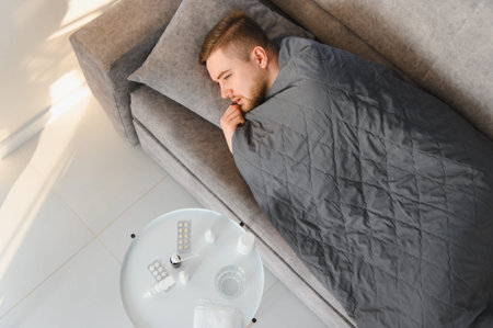 Man catching a cold and flu, staying home and recovering comfortably under a blanket with medical...