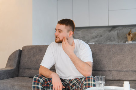 Man experiencing dental pain massaging his cheek while sitting on a couch, needing medication for...