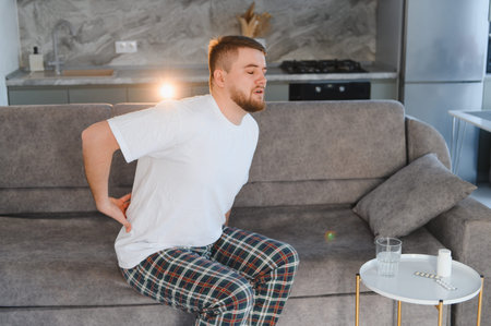 Young man with lower back pain sitting on sofa at home beside medication and water, grimacing from...