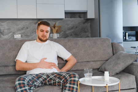 Man struggling with indigestion and abdominal pain, sitting on a sofa with medicine and water nearby