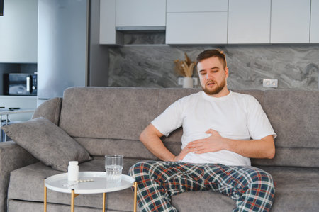 Young man experiencing indigestion and stomach discomfort while sitting on sofa, holding abdomen,...