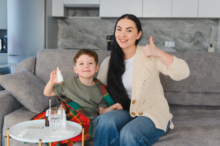 Happy mother and son recovering from cold, sitting on sofa, holding nasal spray and giving thumbs up for healthの写真素材