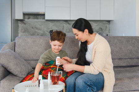 Mom caring for her ill son, giving him liquid medicine to treat symptoms of the flu or cold while resting on a couchの写真素材