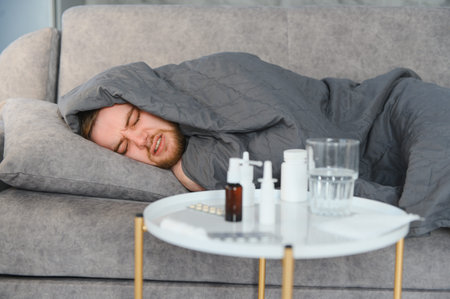 Young man having severe headache and fever, feeling unwell under a gray blanket with medicine on a...