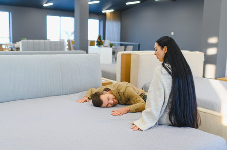Man lying on a mattress while a woman sits, evaluating products in a large furniture and bedding...