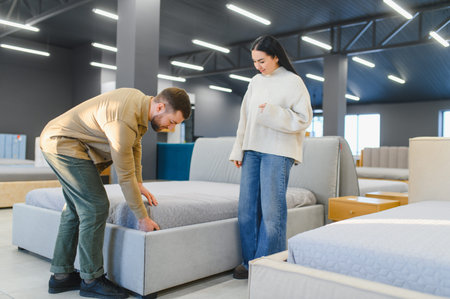 Young couple comparing bed frames and mattresses in a modern furniture showroom, selecting new bedroom furniture for their homeの写真素材