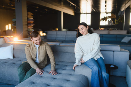 Young couple exploring new sofas in a furniture showroom, feeling the upholstery and choosing...