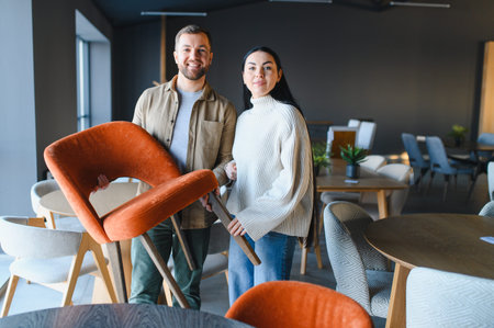 Couple smiling and holding an orange chair in a modern furniture showroom, shopping for home decor and interior designの写真素材