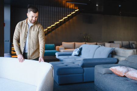 Young man shopping for home furnishings, looking at a white sofa in a modern furniture store with many couchesの写真素材