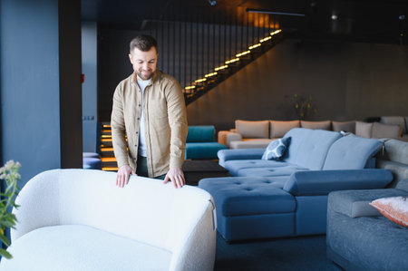Man examining a white sofa, standing in a large furniture showroom with various couches and a staircase in the backgroundの写真素材