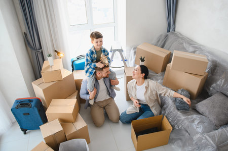Happy family unpacking belongings in their new house. Father carrying child on shoulders, mother...