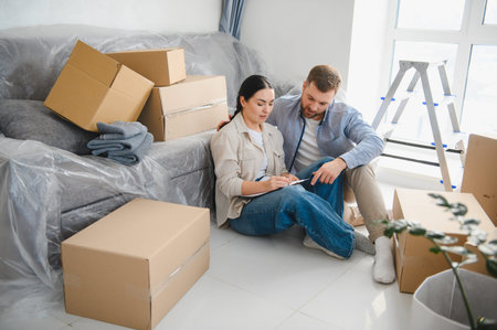 Couple sitting on floor, reviewing checklist for home relocation and packing, surrounded by moving...