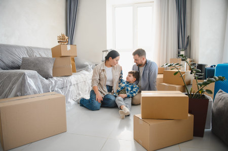 Parents and child sitting on the floor, relaxing among cardboard boxes and packed furniture after...