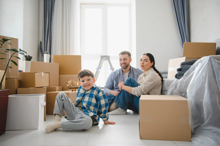 Family including a child, parents sitting on the floor, surrounded by cardboard boxes after moving...