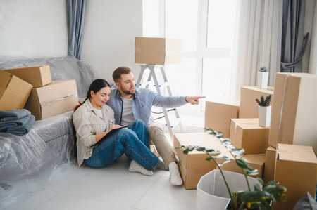 Happy couple sitting on floor among cardboard boxes, discussing design ideas for their new apartment...