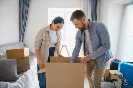 Young couple standing in a new apartment, packing cardboard boxes. They are preparing to move into a...