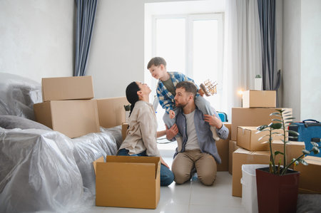 Family with child on dad's shoulders smiling in new home surrounded by cardboard boxes, celebrating...