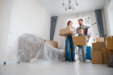 Parents and child moving into new apartment together, carrying cardboard boxes and a plant, ready...
