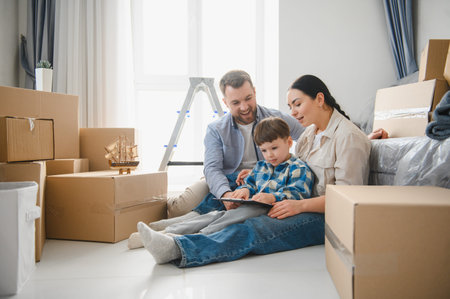 Young family, mother, father and son, sitting on floor in new house surrounded by cardboard boxes,...
