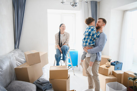 Happy family unpacking belongings in their new apartment, father holding their son while mother sits...