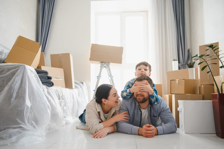 Happy family with child lying on floor surrounded by cardboard boxes, enjoying playful moment in...