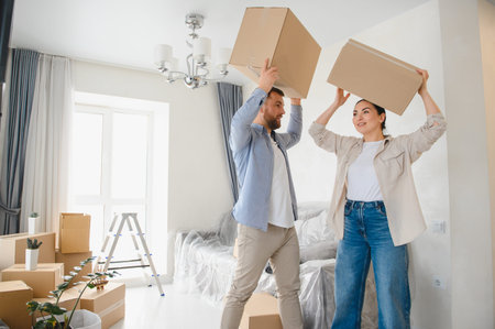 Young couple celebrating new beginnings while carrying cardboard boxes in their new home, surrounded...