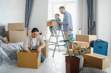 Happy family relocating, parents and child packing cardboard boxes together in a bright room,...