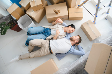 Happy couple celebrating home ownership, smiling and resting on the floor among cardboard boxes in...