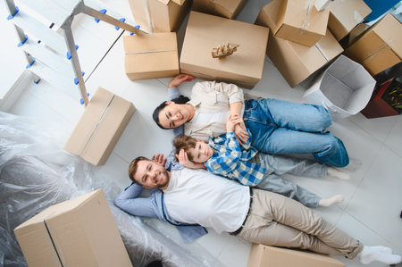Happy family with a child lying on the floor surrounded by moving boxes in their new home, feeling...