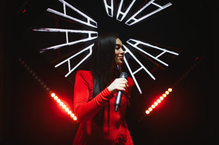 Female singer in a red dress performing a song on stage under bright concert lights and abstract led screen graphicsの写真素材