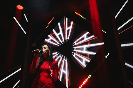 Female performer singing on stage, holding a microphone. Stage lighting and led display creating a...