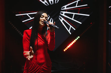 Female singer in a red dress performing on a dark stage, holding a microphone, with glowing lights in the backgroundの写真素材